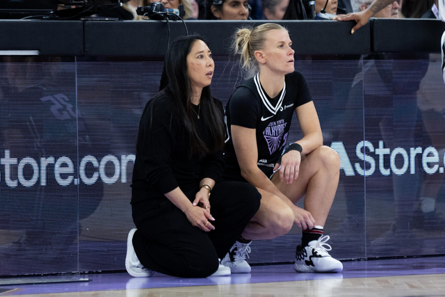 Two women are crouched by a basketball court. One wears a black tracksuit, and the other wears a sports jersey. Both appear focused on the game.